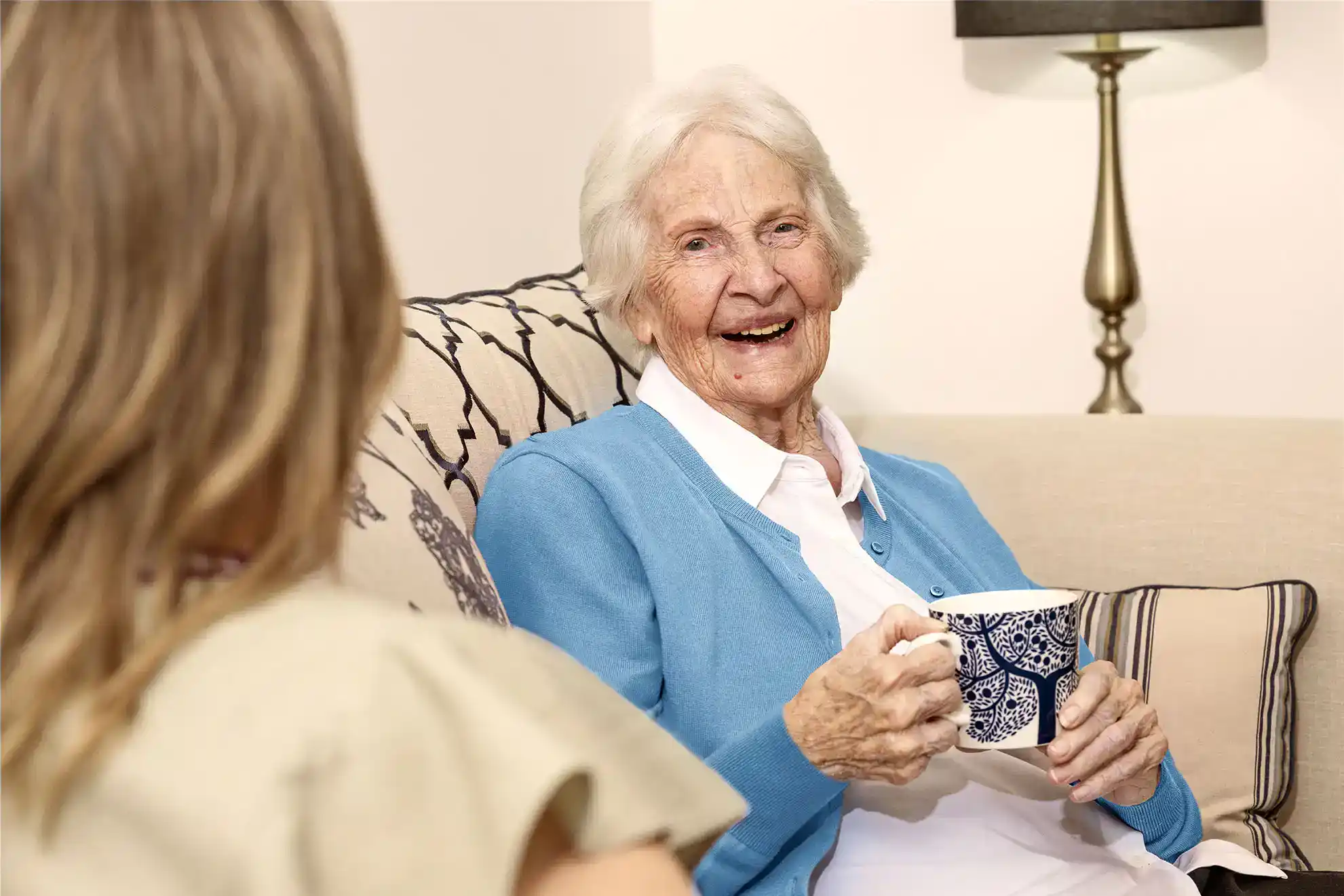 Elderly woman smiling and holding a cup of tea while chatting with another person in a comfortable living room.
