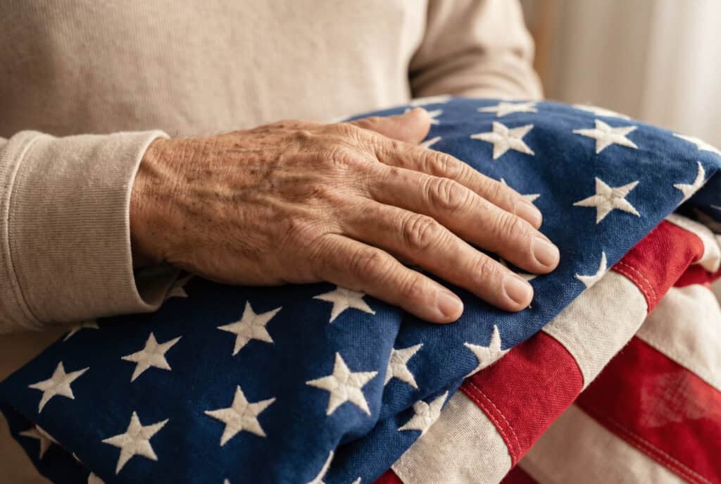 Close-up of an elderly person’s hand resting on a folded American flag, highlighting themes of military service, remembrance, and honor.