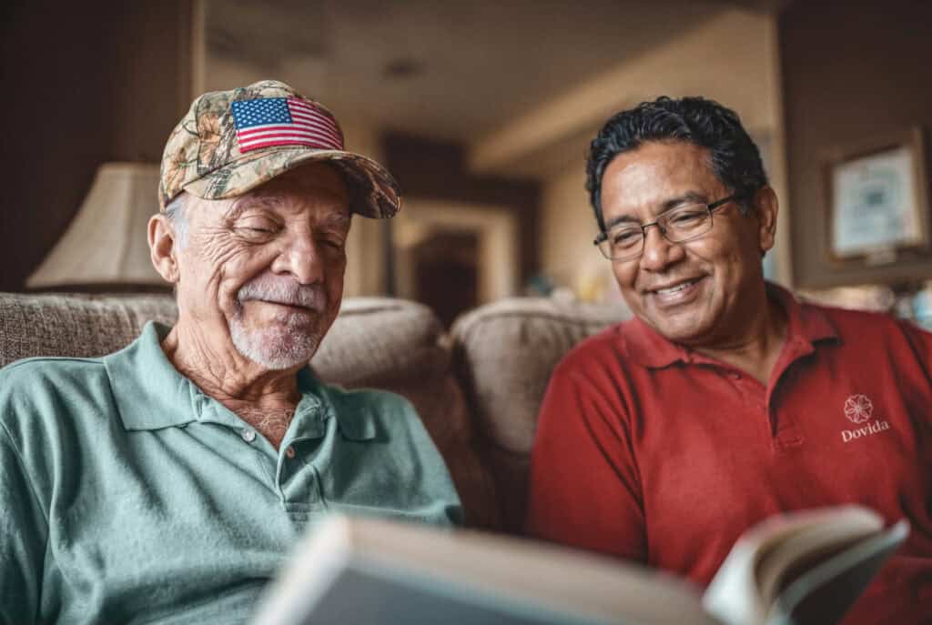 A veteran in a camouflage American flag cap sits indoors reading a book alongside a smiling caregiver in a red Dovida polo shirt.