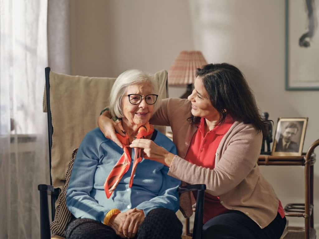 Dovida Caregiver helping elderly woman adjust scarf while seated at home