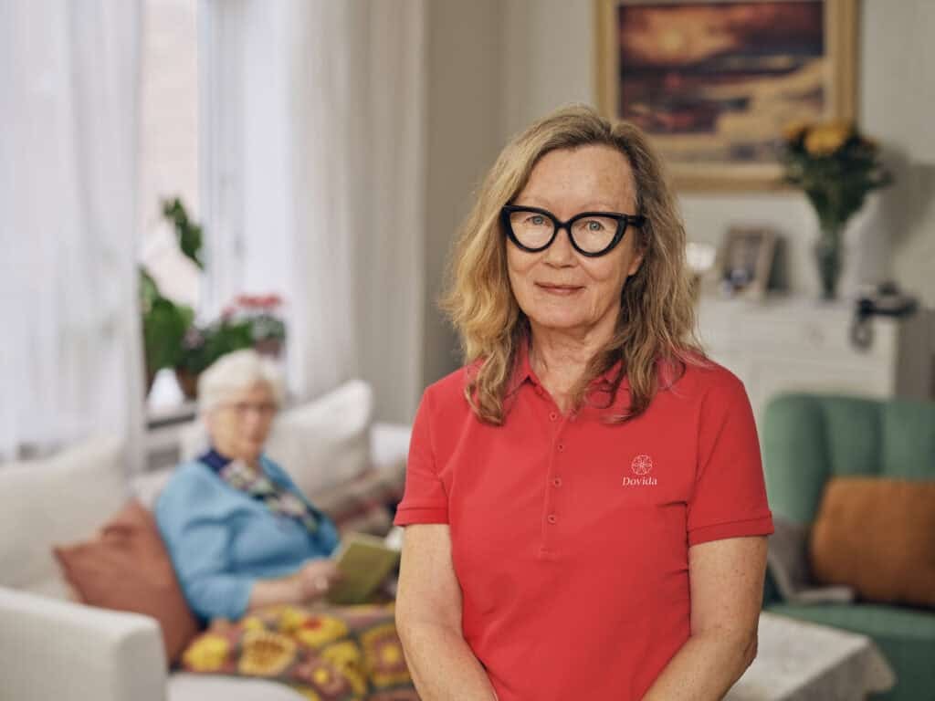 Female Dovida caregiver in red uniform standing in a living room with an elderly woman seated behind him.
