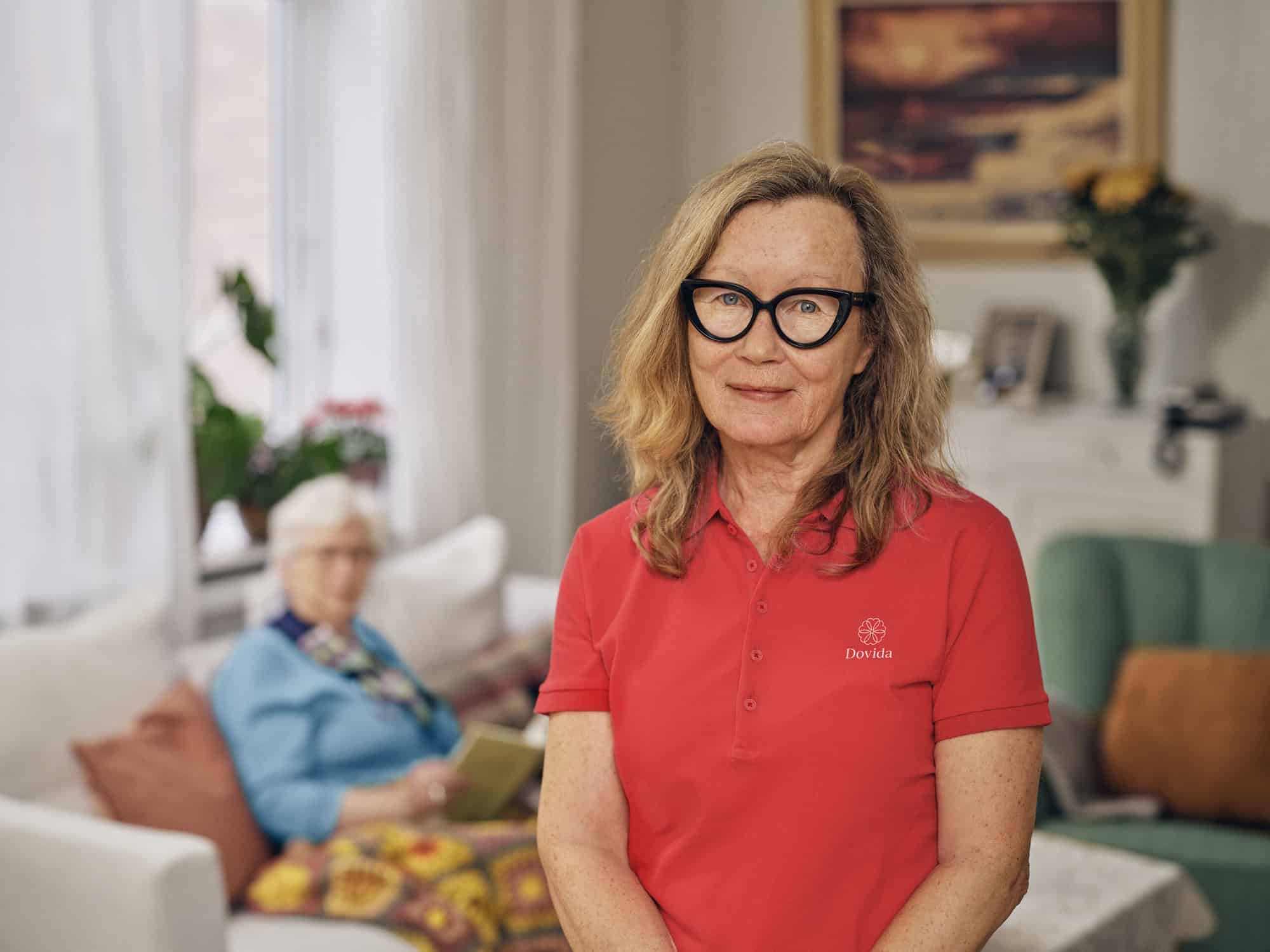 Female Dovida caregiver in red uniform standing in a living room with an elderly woman seated behind him.