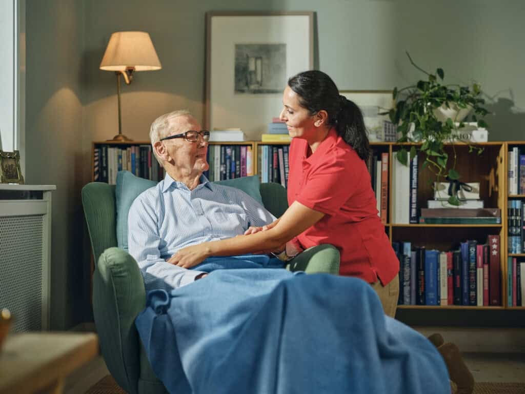 A Dovida Caregiver supports an elderly man in a chair, reflecting the dedication found in caregiver training and Dovida jobs.
