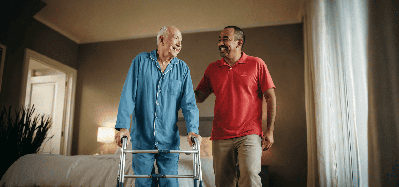 Caregiver supporting elderly man using a walking frame in bedroom at home