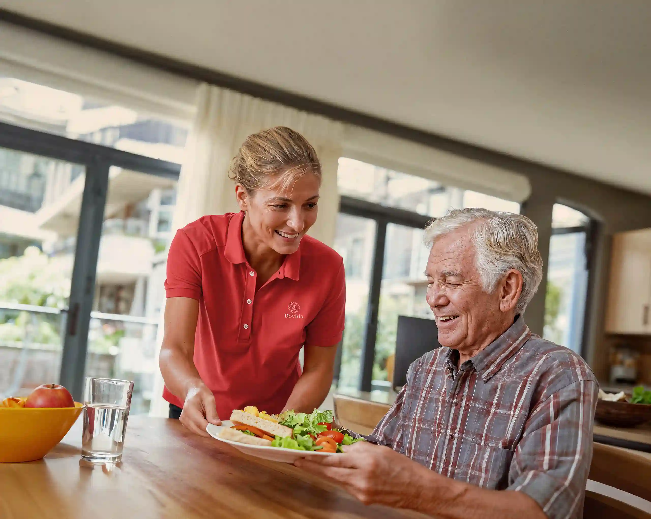 Caregiver serving a healthy meal to an elderly man at a dining table.
